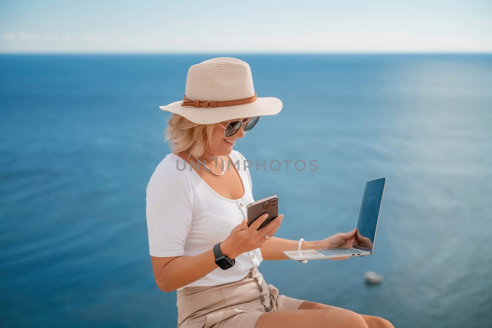 Freelance women sea working on the computer. Good looking middle aged woman typing on a laptop keyboard outdoors with a beautiful sea view. The concept of remote work. by Matiunina