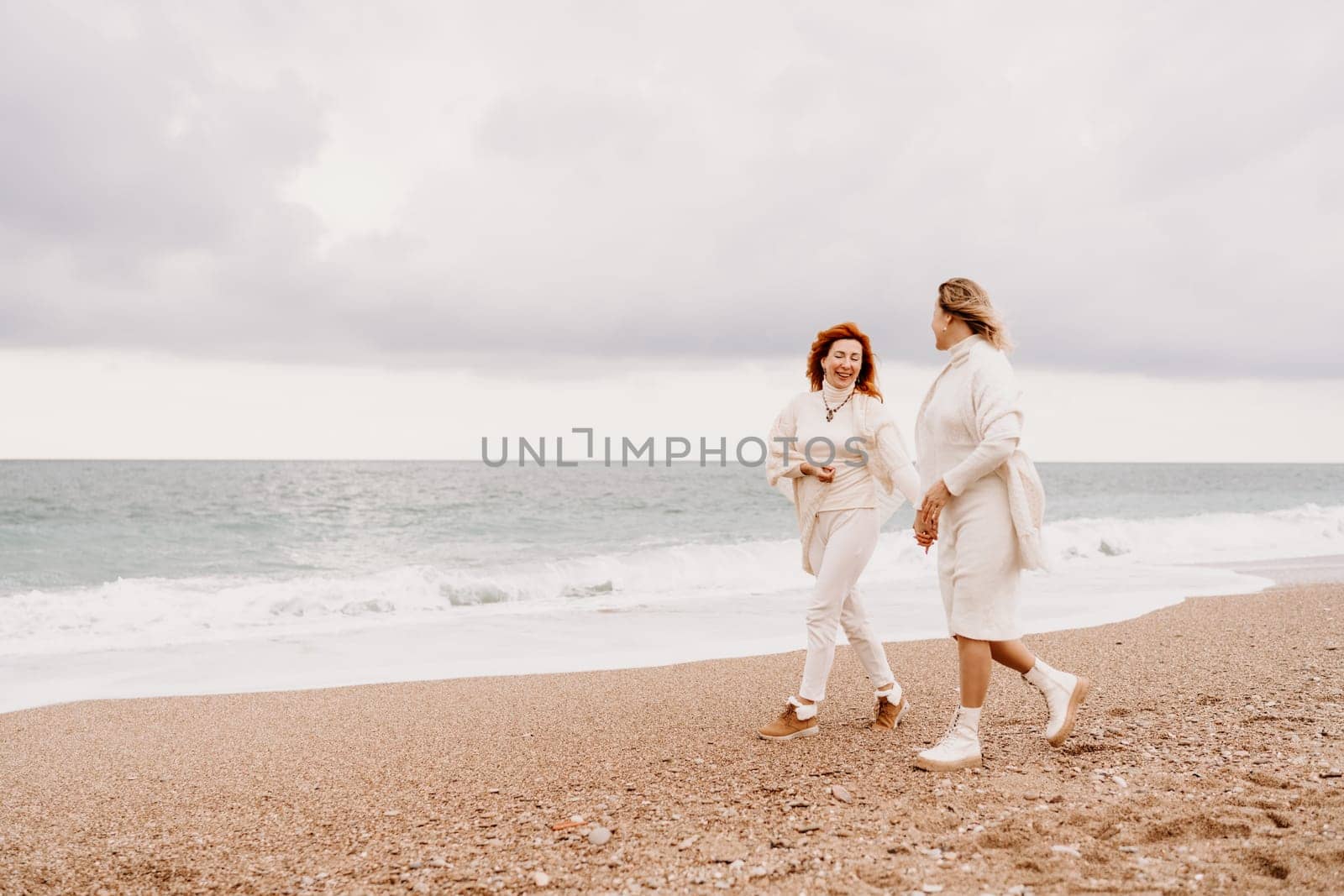 Women sea walk friendship spring. Two girlfriends, redhead and blonde, middle-aged walk along the sandy beach of the sea, dressed in white clothes. Against the backdrop of a cloudy sky and the winter sea. Weekend concept