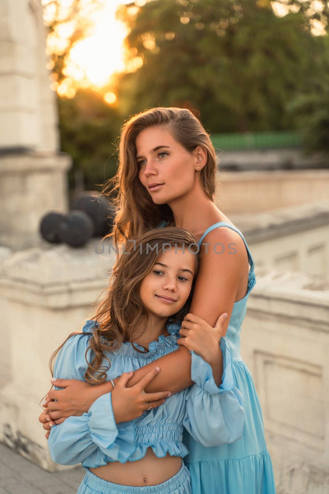 Mother aughter sunset. in blue dresses with flowing long hair against the backdrop of sunset. The woman hugs and presses the girl to her