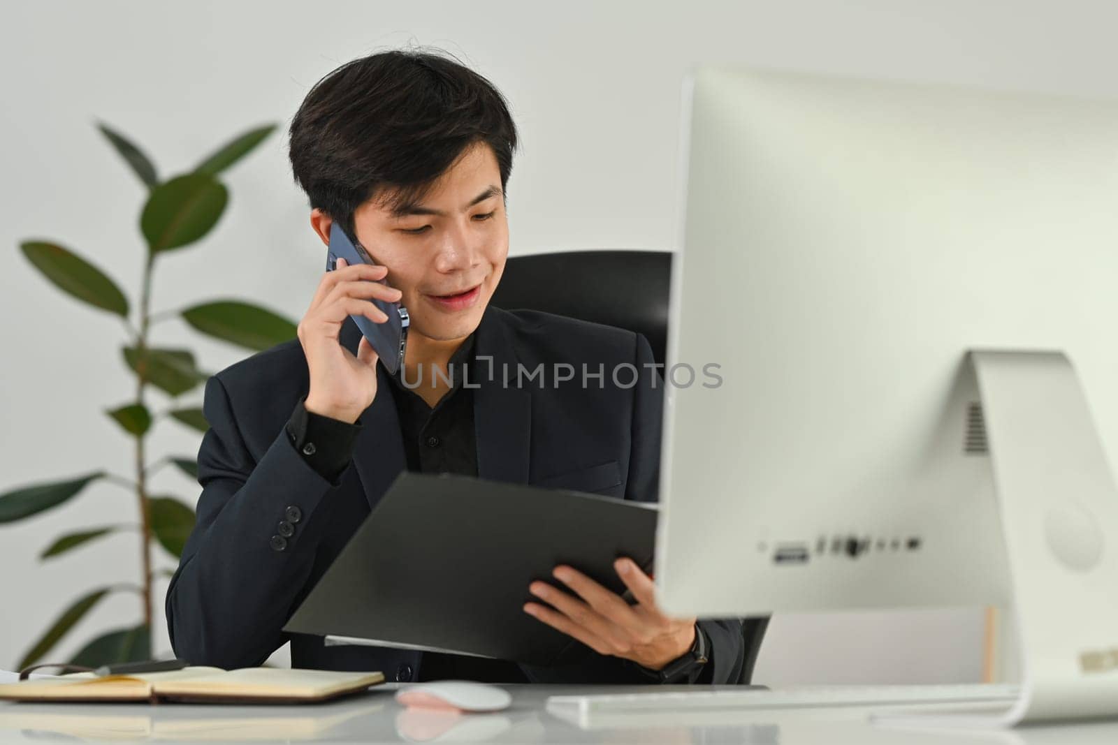 Smiling male accountant advisor holding open office binder and talking on mobile phone while sitting at workstation by prathanchorruangsak