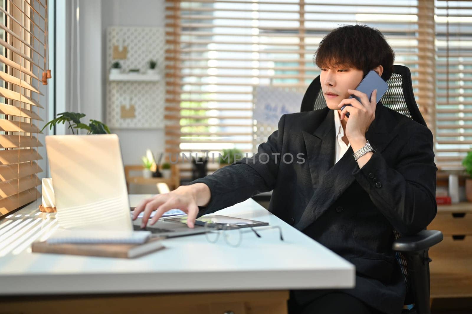 Young asian male business executive looking at laptop screen, discussing project on mobile phone.