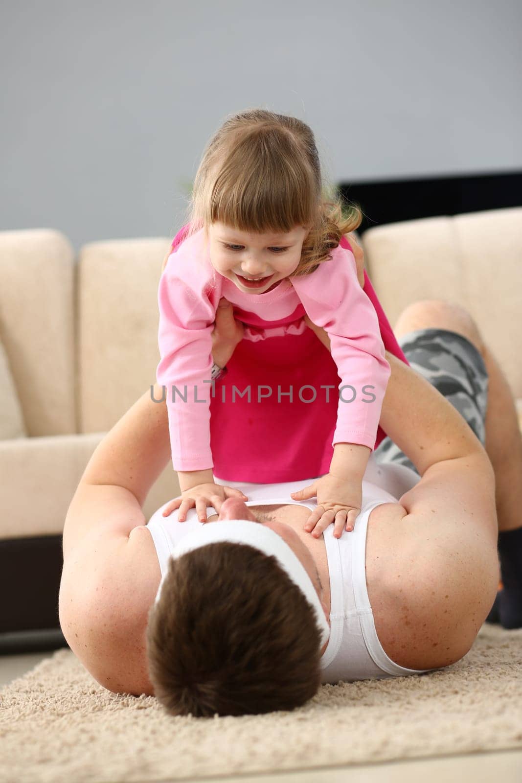 Joyful young father man lying on carpet floor lifting excited happy little daughter at home. Carefree family having fun practicing acroyoga as couple in the living room