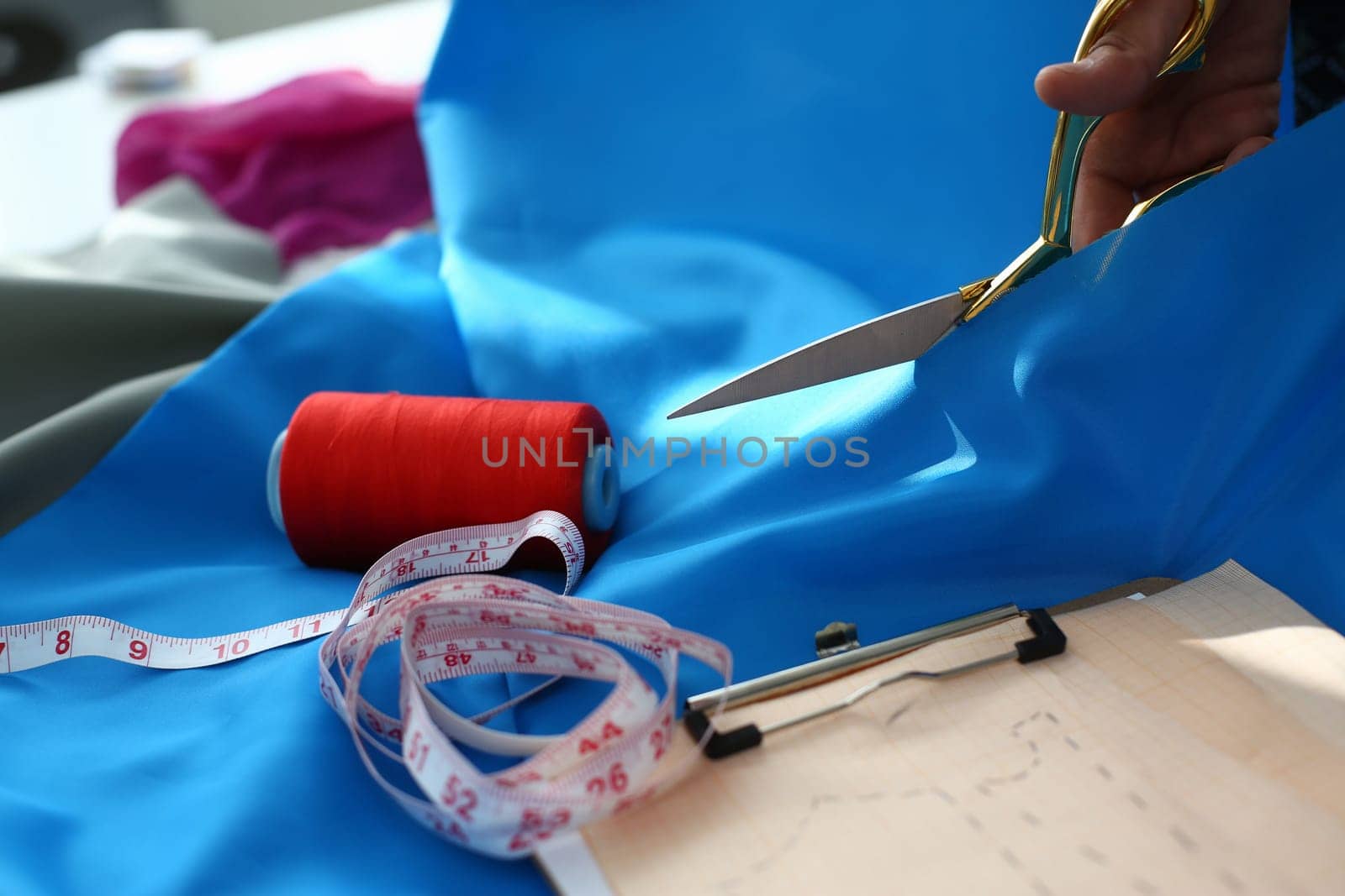 Tailor cuts piece of blue fabric at table in fashion studio. Thread measuring tape and tailoring concept