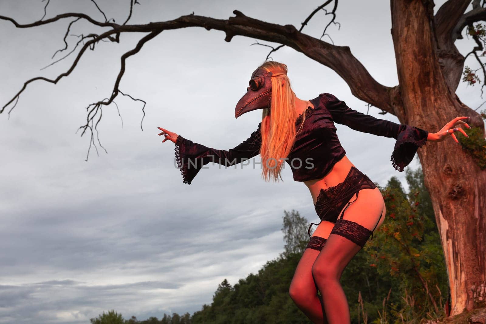 Blonde in crow mask poses in nature near old tree by palinchak