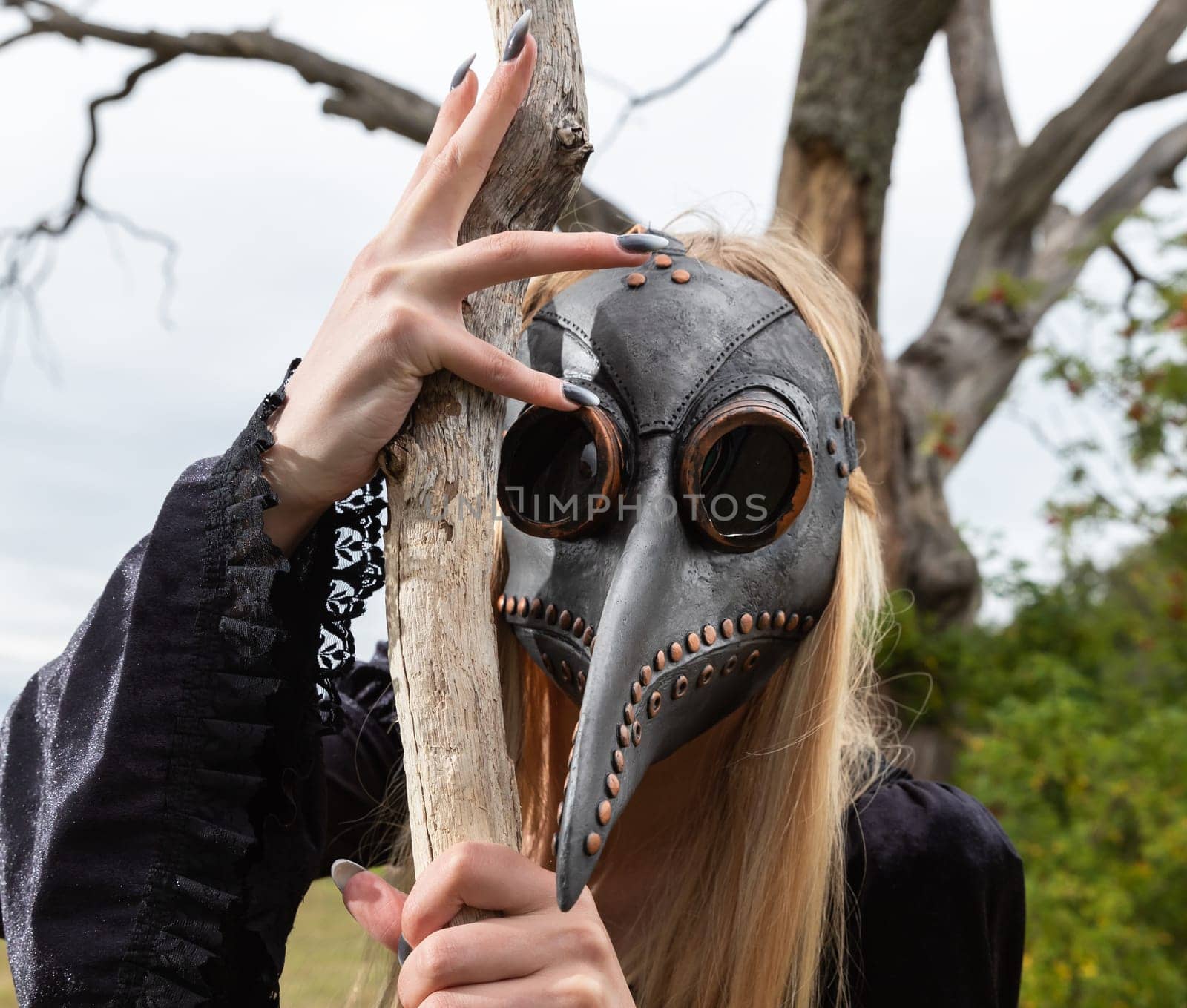 Blonde in crow mask poses in nature near old tree by palinchak