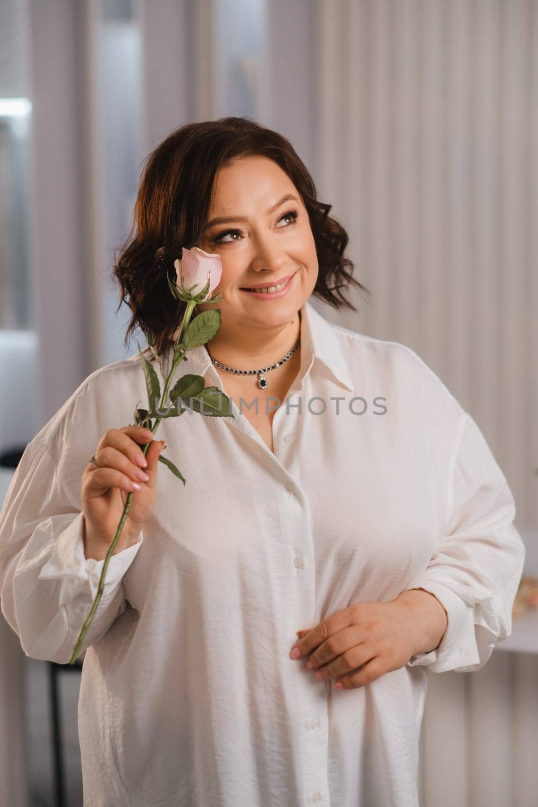 An adult woman in a white shirt holds one rose in her hands while standing in the interior. Pink roses.