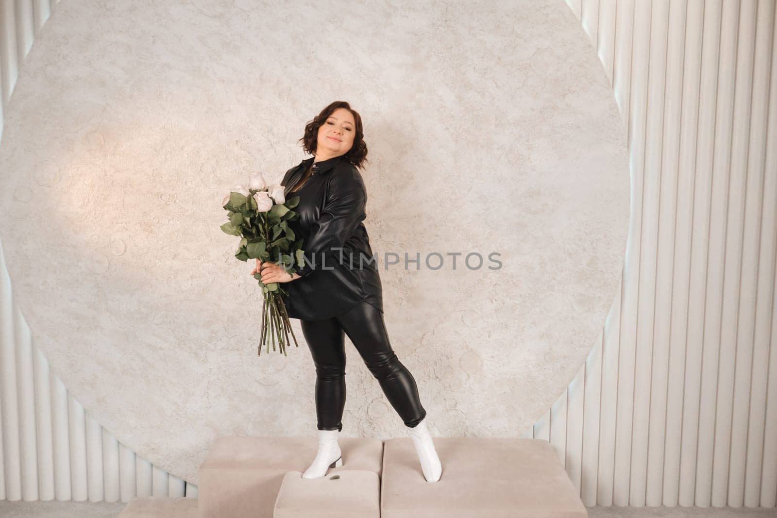 a stylish adult woman in black leather clothes stands with a bouquet of pink roses in the interior.