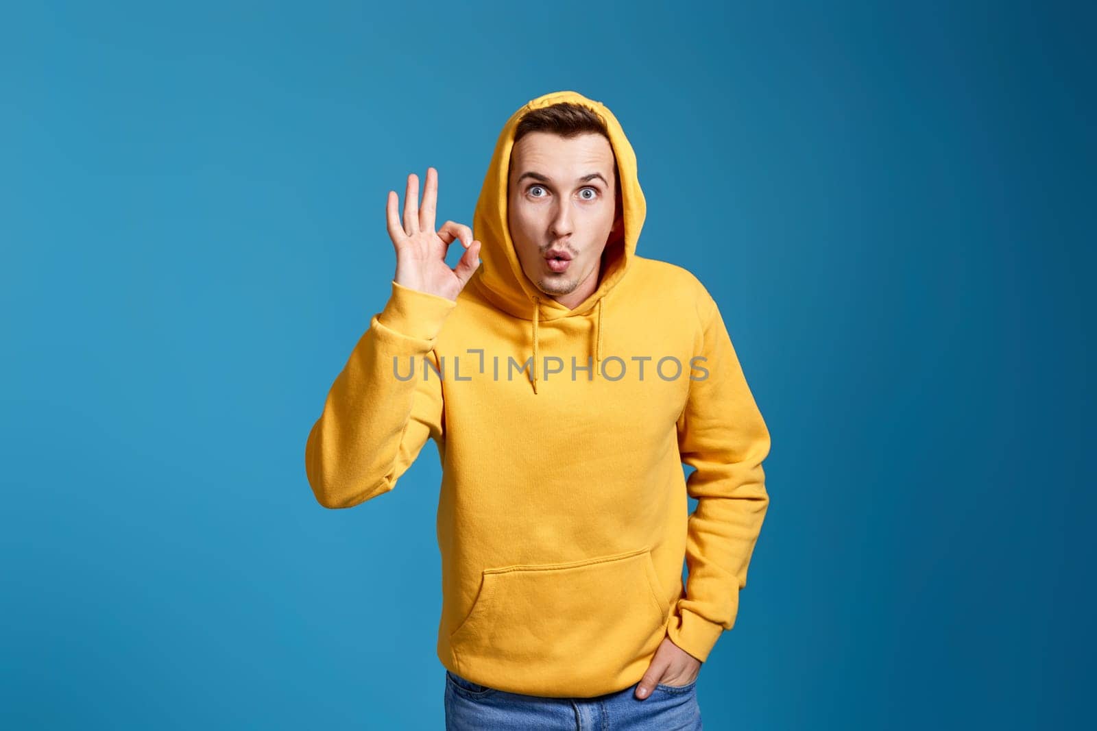 happy young man in yellow sweatshirt showing Ok gesture isolated on blue background.
