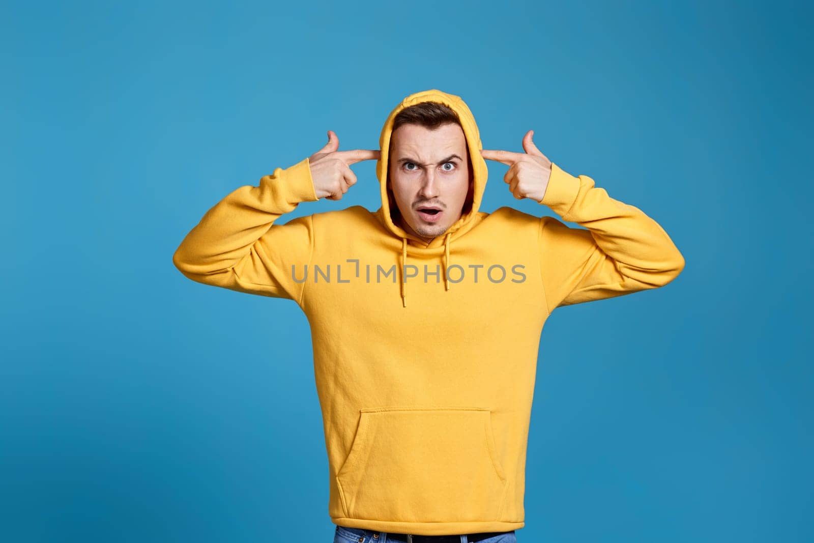 shocked man in yellow sweatshirt holding his fingers on his temples isolated on blue background
