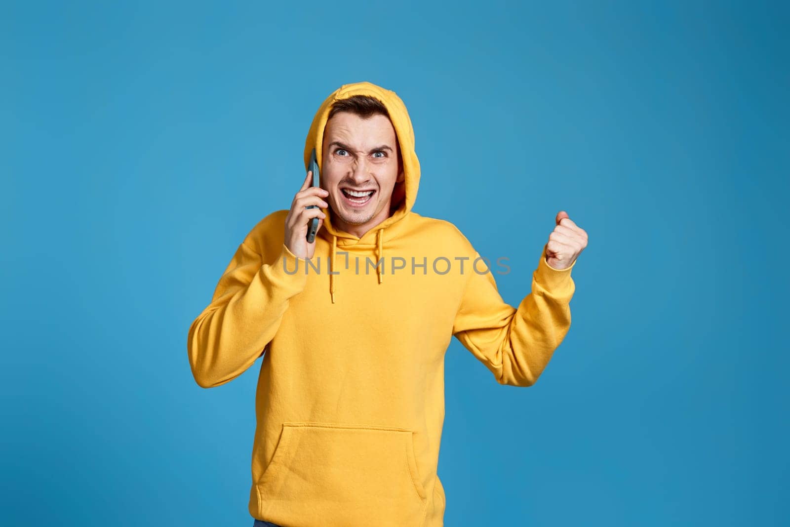 Upset angry man in yellow sweatshirt holding mobile phone and feel outraged isolated on blue background