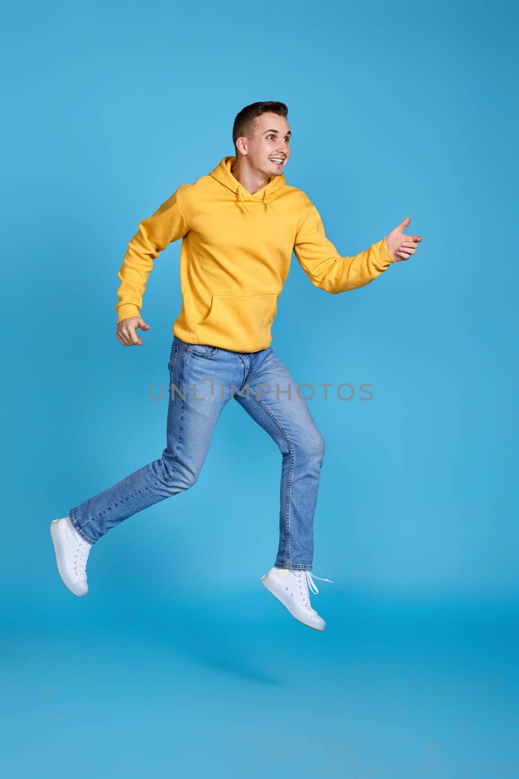very excited happy man in yellow sweatshirt and jeans jumping isolated on blue background. Full length