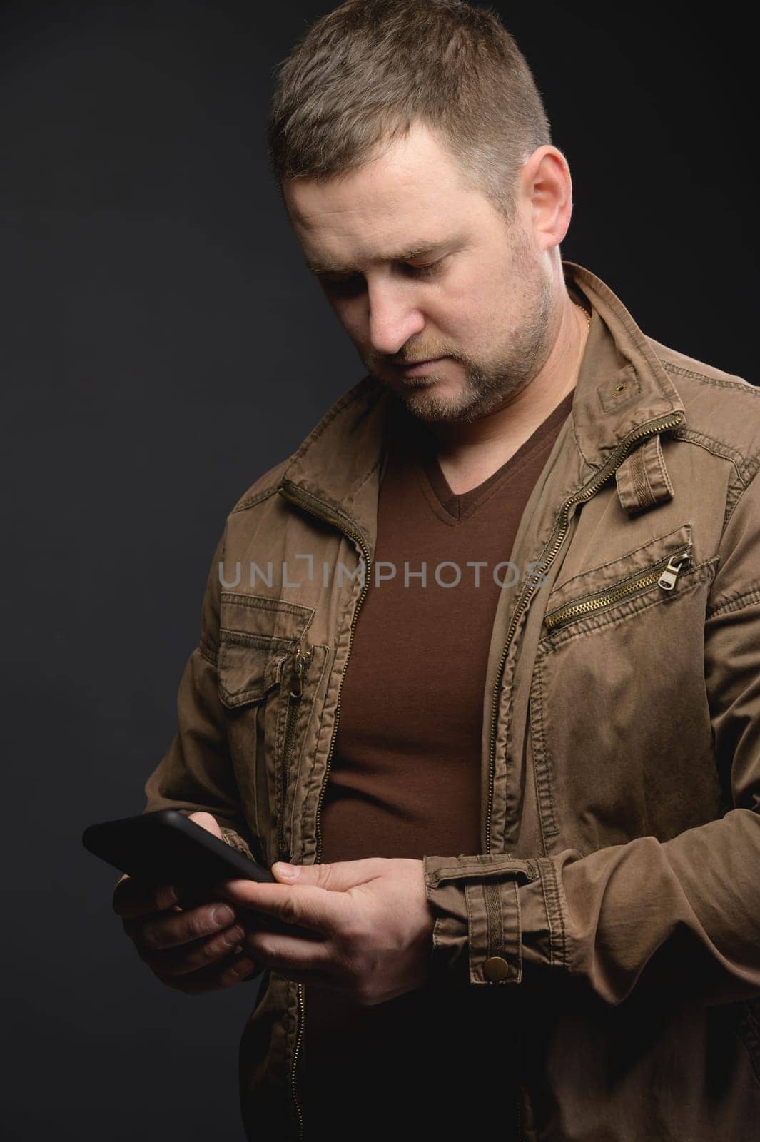 Portrait of a confident stylish young man in casual clothes holding a mobile phone, looking at the screen, standing against a black background. Successful businessman. Business, technology.
