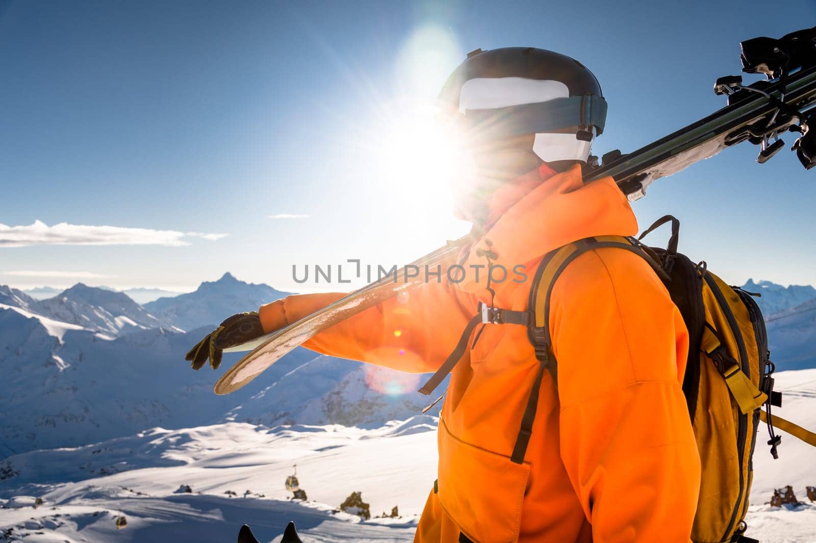 Portrait of a man holding skis on his shoulder and looking at a snowy mountain against the backdrop of a shining sun, his face is not visible, an unrecognizable person by yanik88