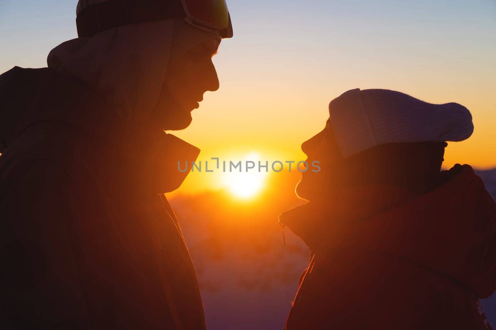 Side view of a silhouetted couple of hikers standing together against a beautiful sunrise or sunset on top of a mountain with a clear sky in the background. Man and woman look at each other.