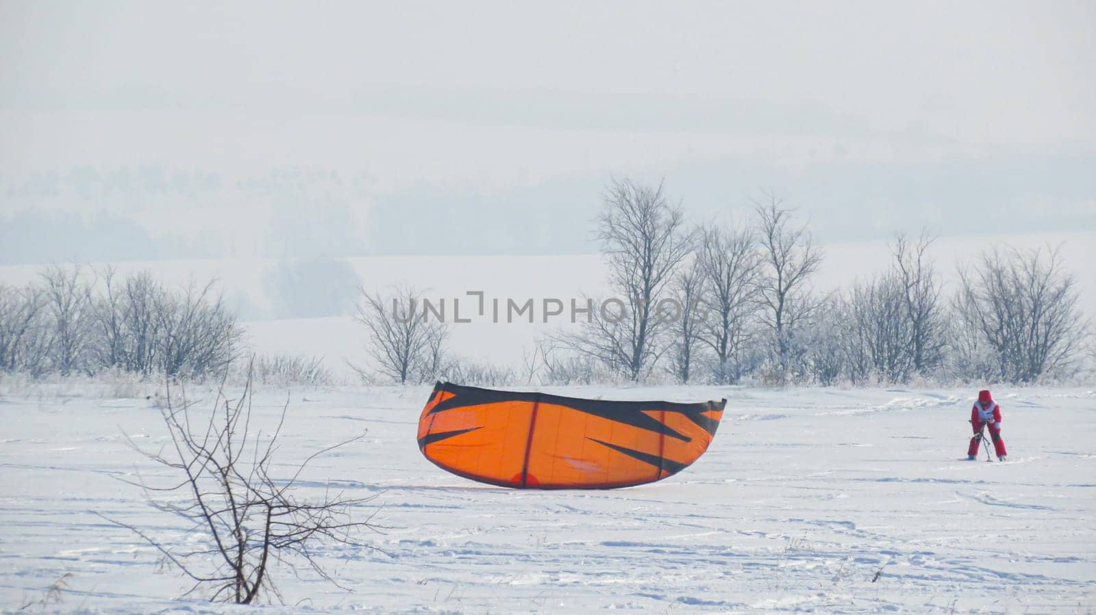 A girl in a red jumpsuit, snowkiting, parachuting in the field in winter. Lying kiting. Girl is kiting in the field.