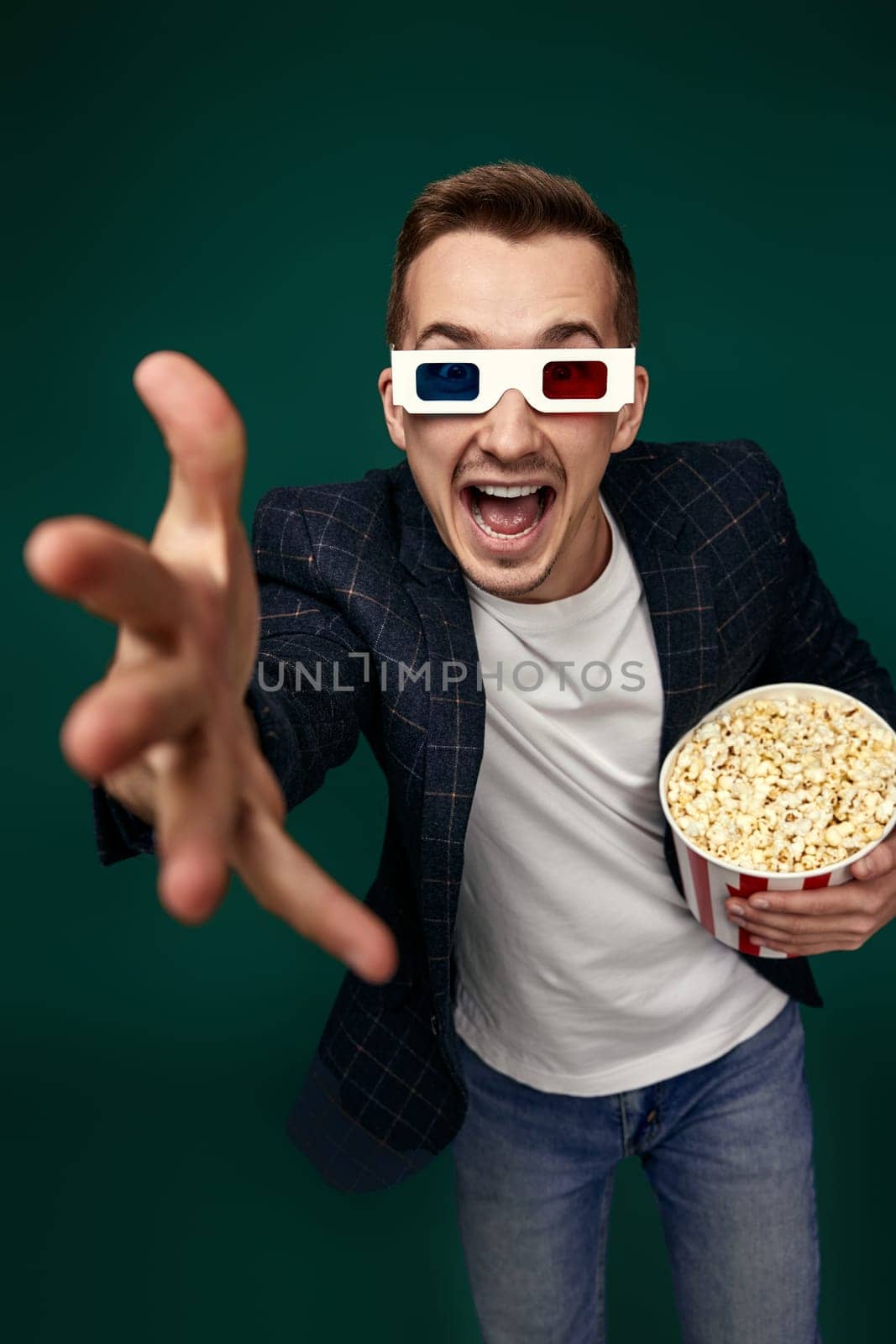 happy caucasian man in 3d glasses holding bucket of popcorns and pointing at camera on green background