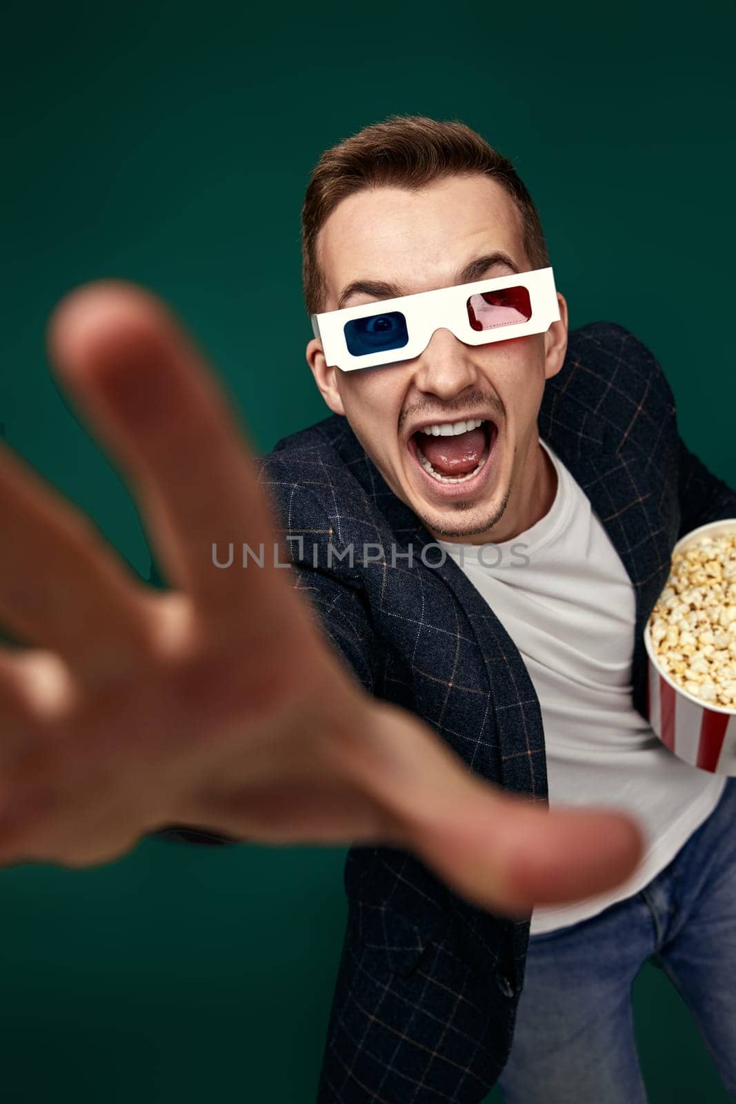 happy caucasian man in 3d glasses holding bucket of popcorns and pointing at camera on green background
