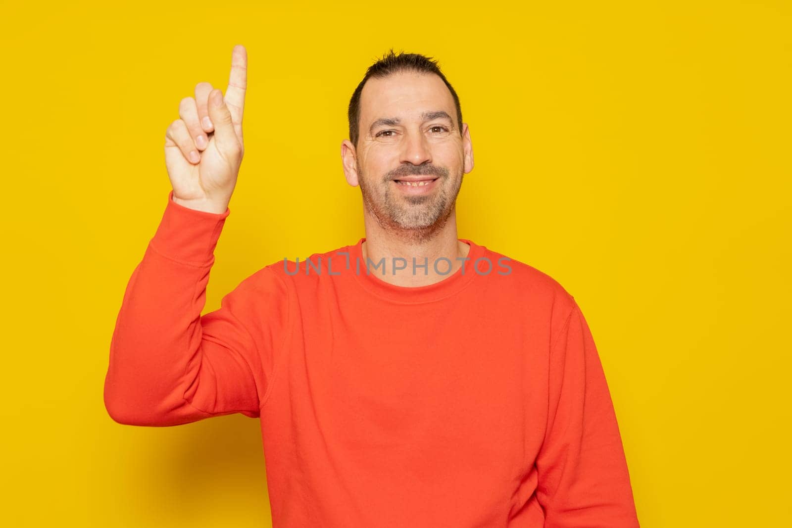 Handsome hispanic man with a beard in a red sweatshirt raising his index finger excited, he has had a great idea and he wants to tell everyone about it. Isolated on yellow background