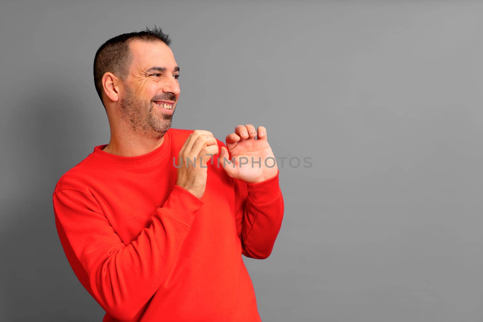 Hispanic man in his 40s wearing a red sweater poses embarrassed to the side with his hands in a defensive position, isolated on gray background