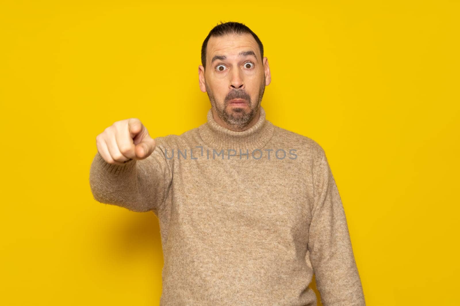 Bearded hispanic man in his 40s wearing a beige turtleneck pointing at something scary, he is very scared and feeling totally terrified. Isolated on yellow background