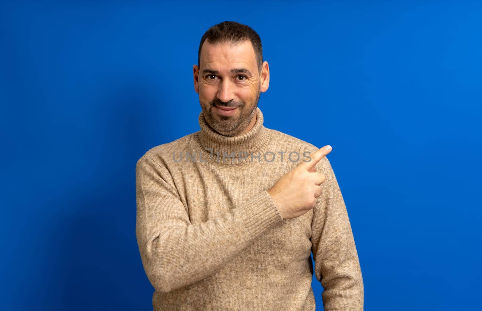 Friendly bearded Hispanic man in his 40s wearing a beige turtleneck pointing to the side, isolated over a blue background by Barriolo82