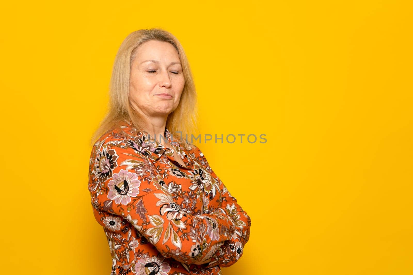Caucasian blonde woman in her 40s wearing a patterned dress posing funny with her arms crossed and her eyes closed, isolated over yellow background