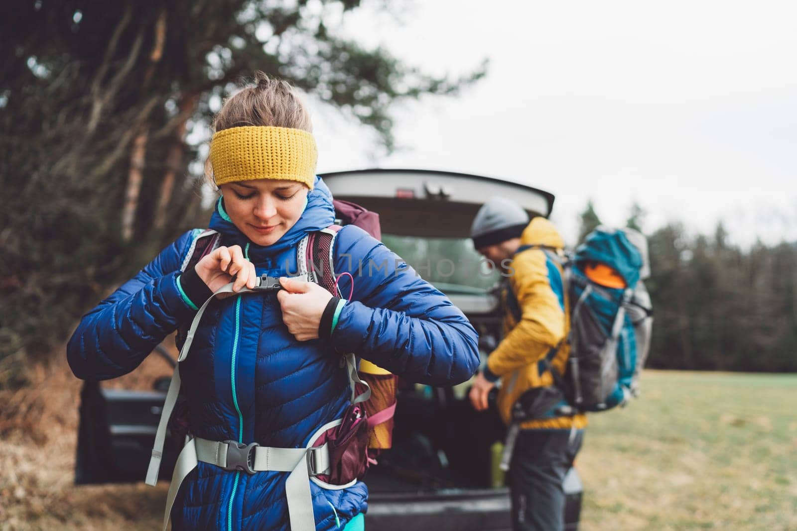 Woman hiker putting on her backpack and her partner in the background taking things out of the car trunk by VisualProductions