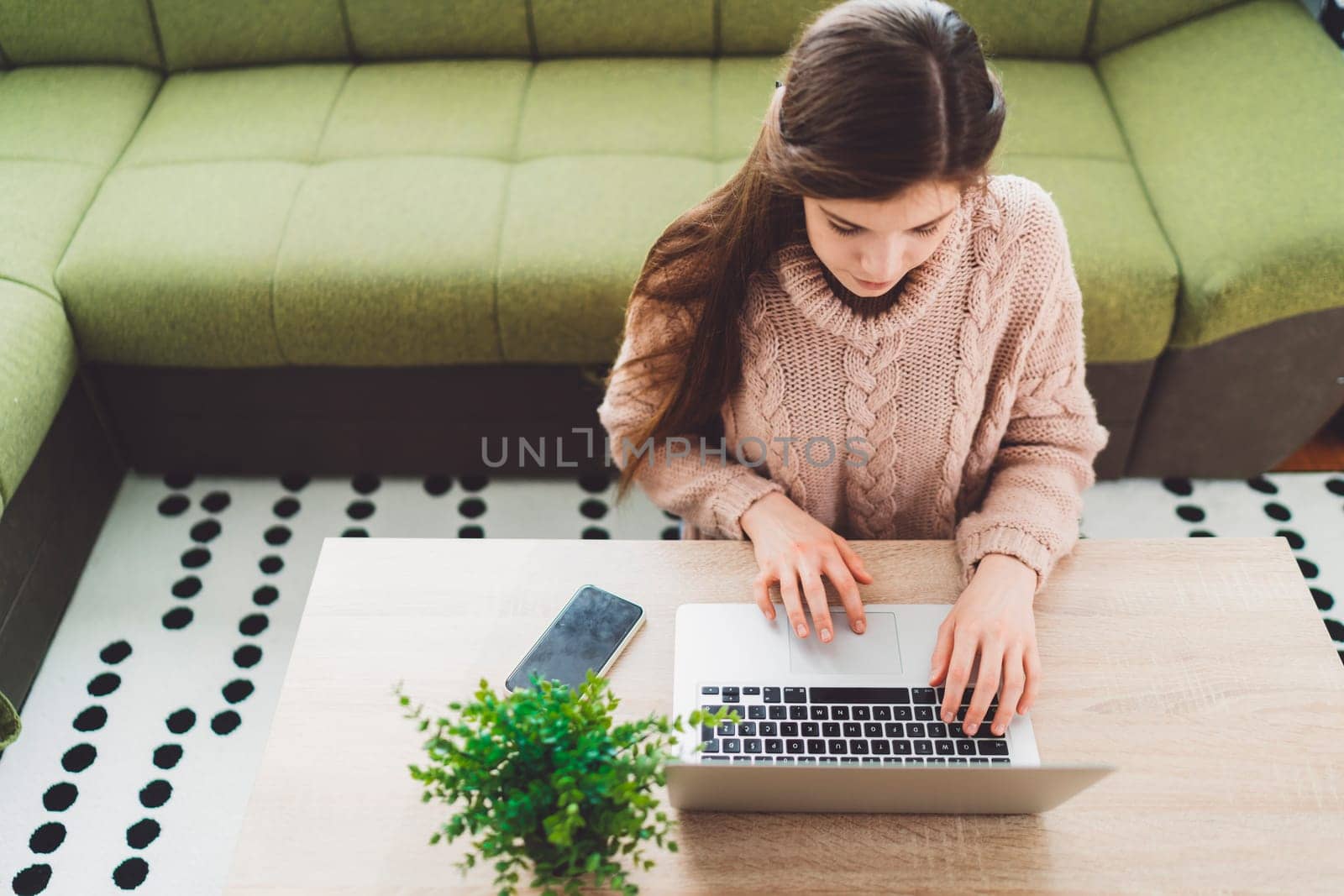 Young caucasian woman with long brown hair in pastel pink sweater and jeans working from home on her laptop and phone. Cheerful woman student studying at home.