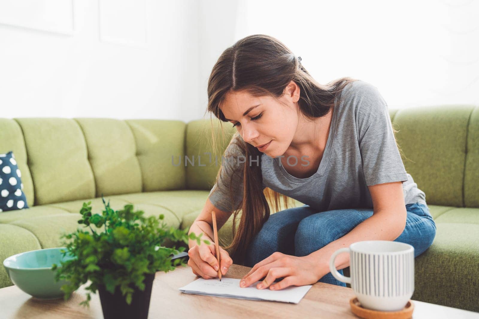 Woman leaning down on the coffee table writing an address on the letter she has to send by VisualProductions