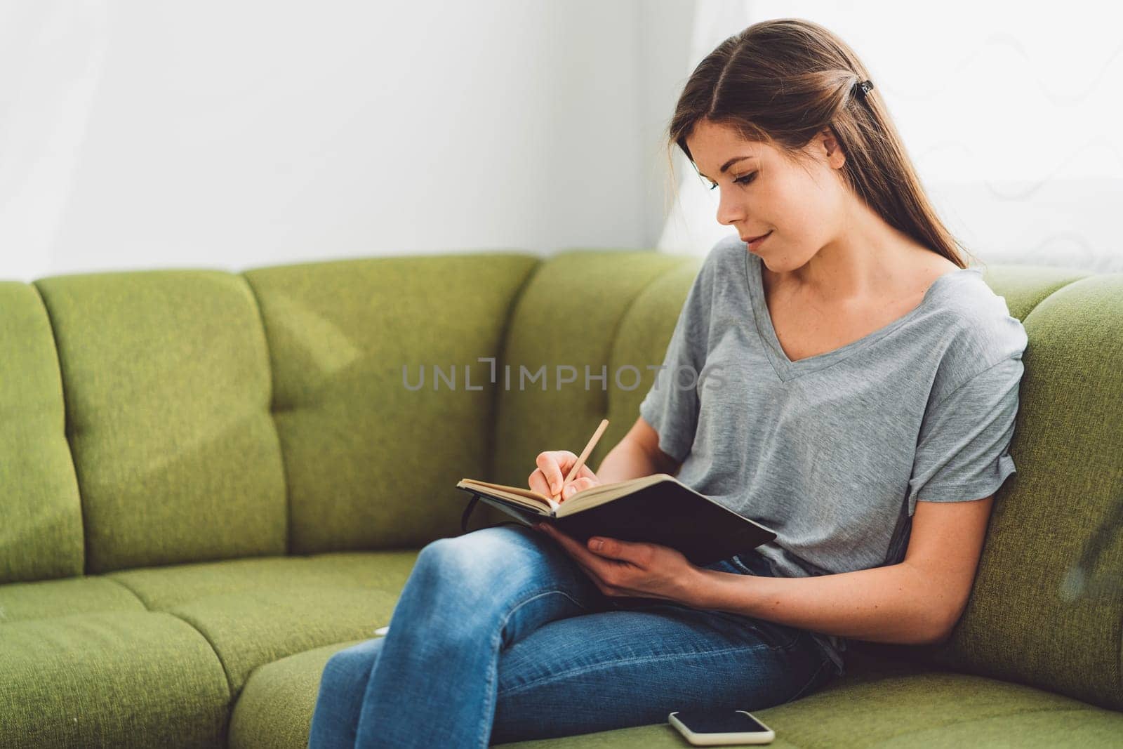 Young caucasian woman sitting on the green couch writing in her notebook by VisualProductions