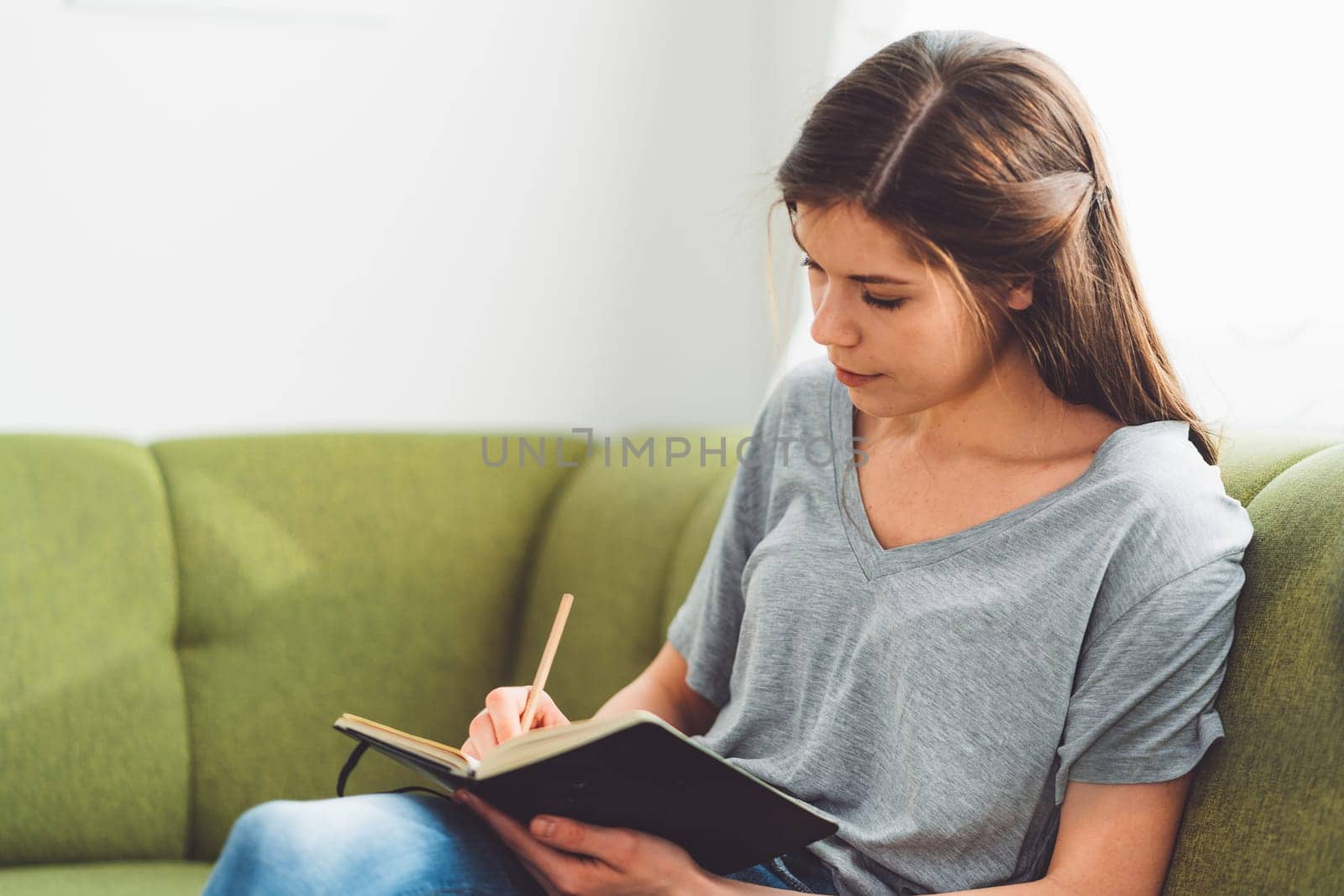 Young caucasian woman with long brown hair in jeans working from home, writing notes. Cheerful woman student studying at home.