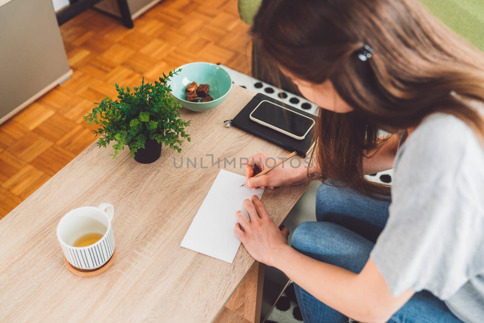 Young caucasian woman with long brown hair in jeans working from home, writing notes. Cheerful woman student studying at home.
