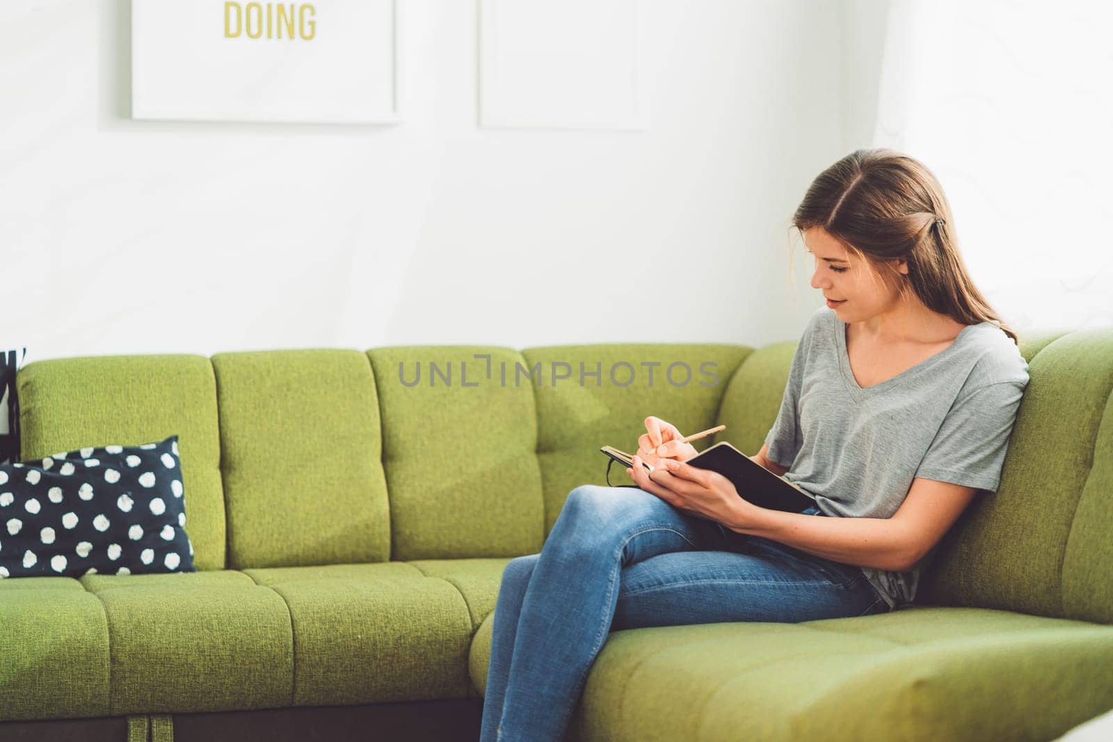 Relaxed young woman writing in her notebook, sitting on the green couch by VisualProductions