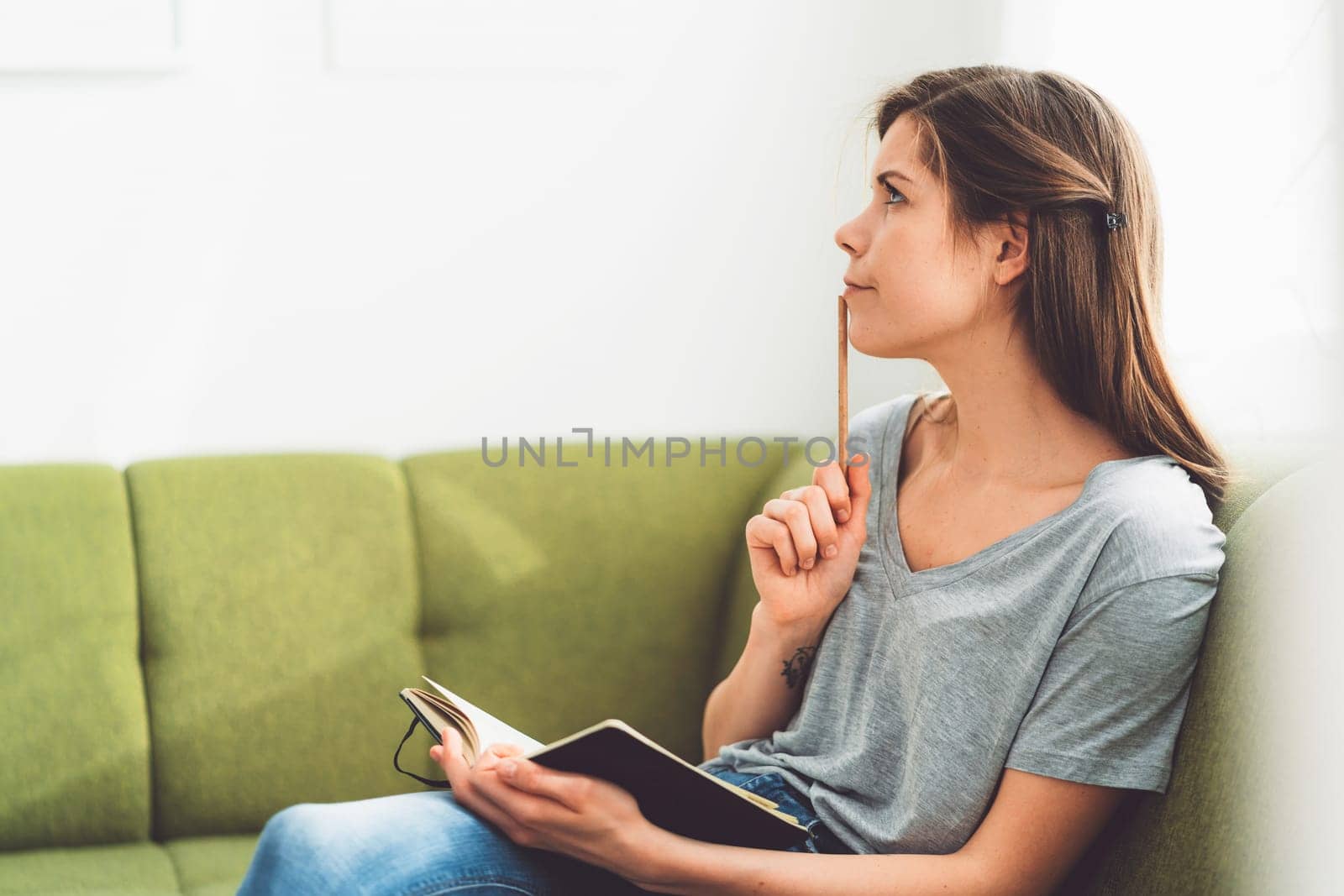Young woman holding a pencil next to her mouth writing ideas down in her notebook by VisualProductions