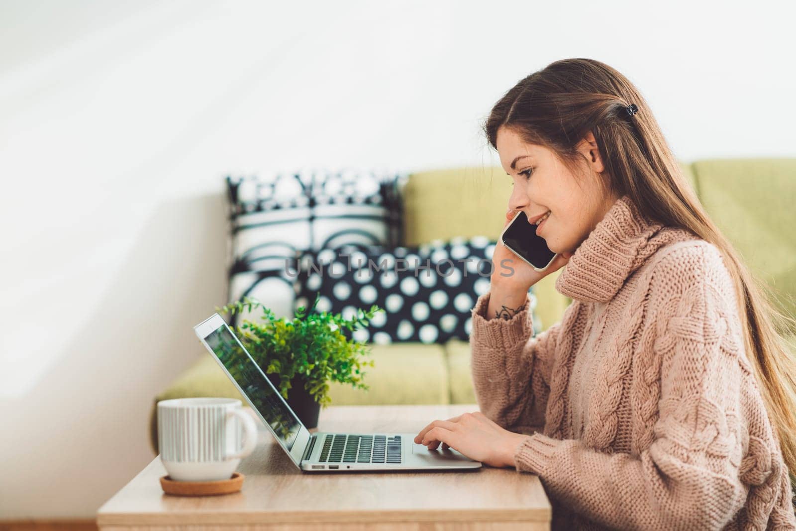 Young caucasian woman with long brown hair in pastel pink sweater and jeans working from home on her laptop and phone. Cheerful woman student studying at home.
