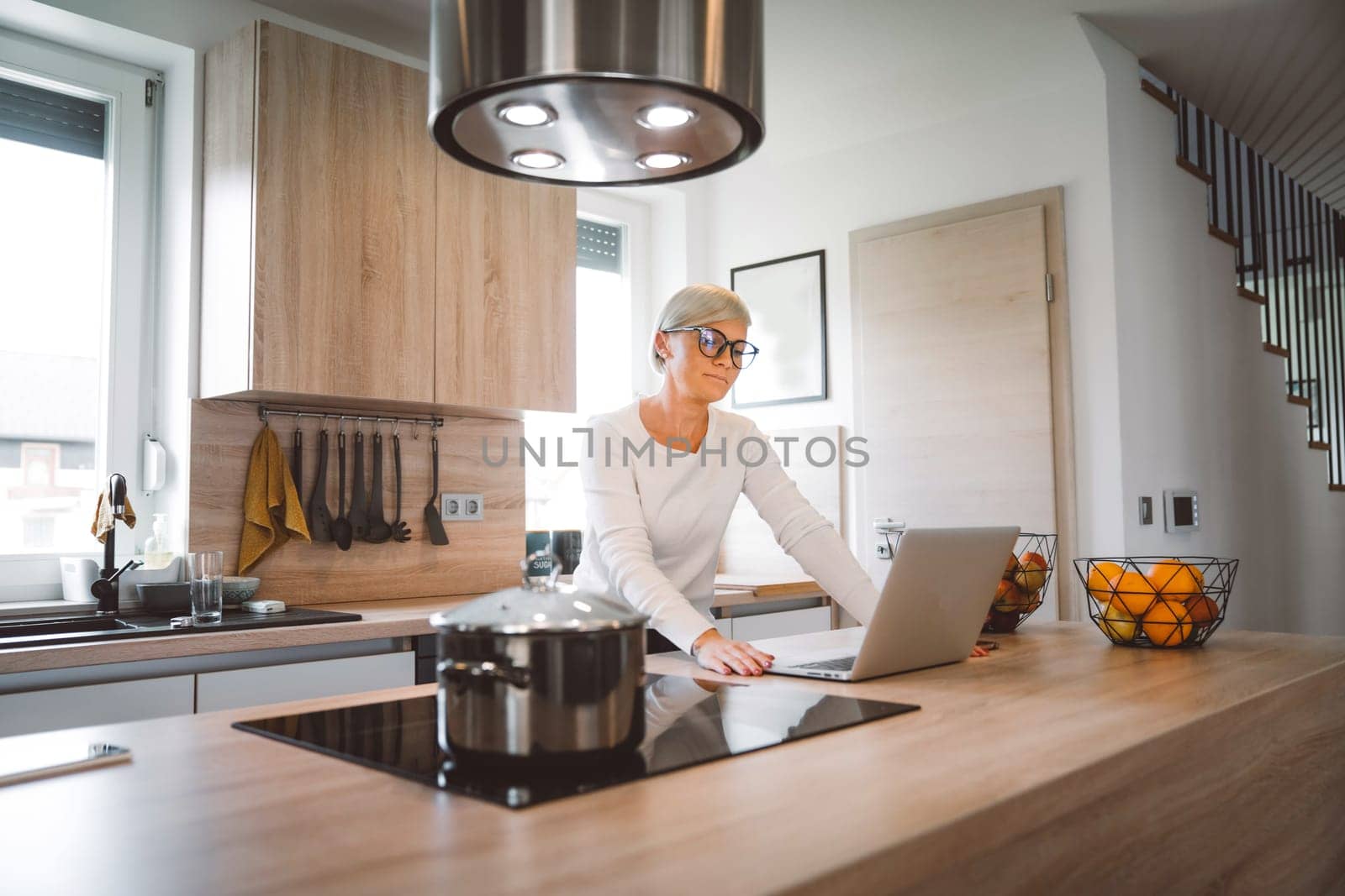 Blonde caucasian woman working in the kitchen on her laptop by VisualProductions