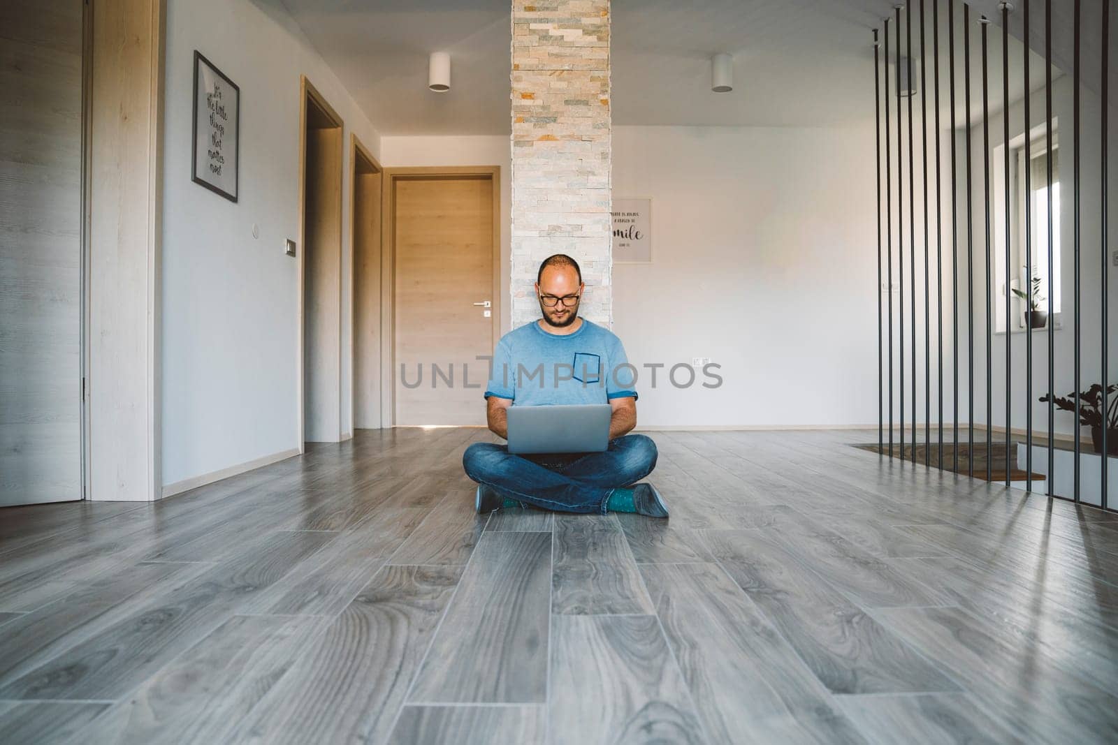 Man sitting on the hallway with his legs crossed working on his laptop from home by VisualProductions