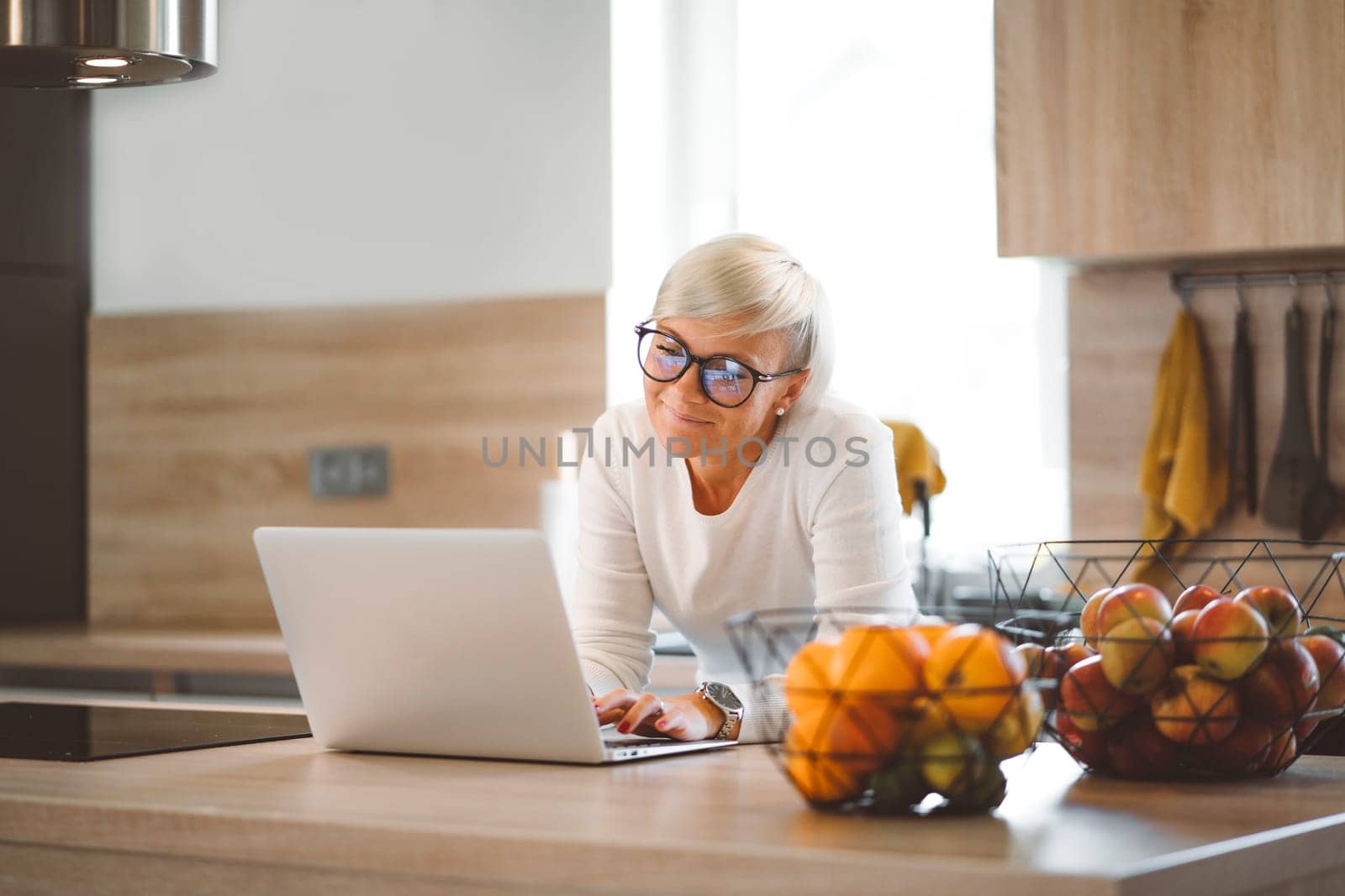 Blonde caucasian woman working on her laptop while leaning on the kitchen counter about to cook lunch by VisualProductions