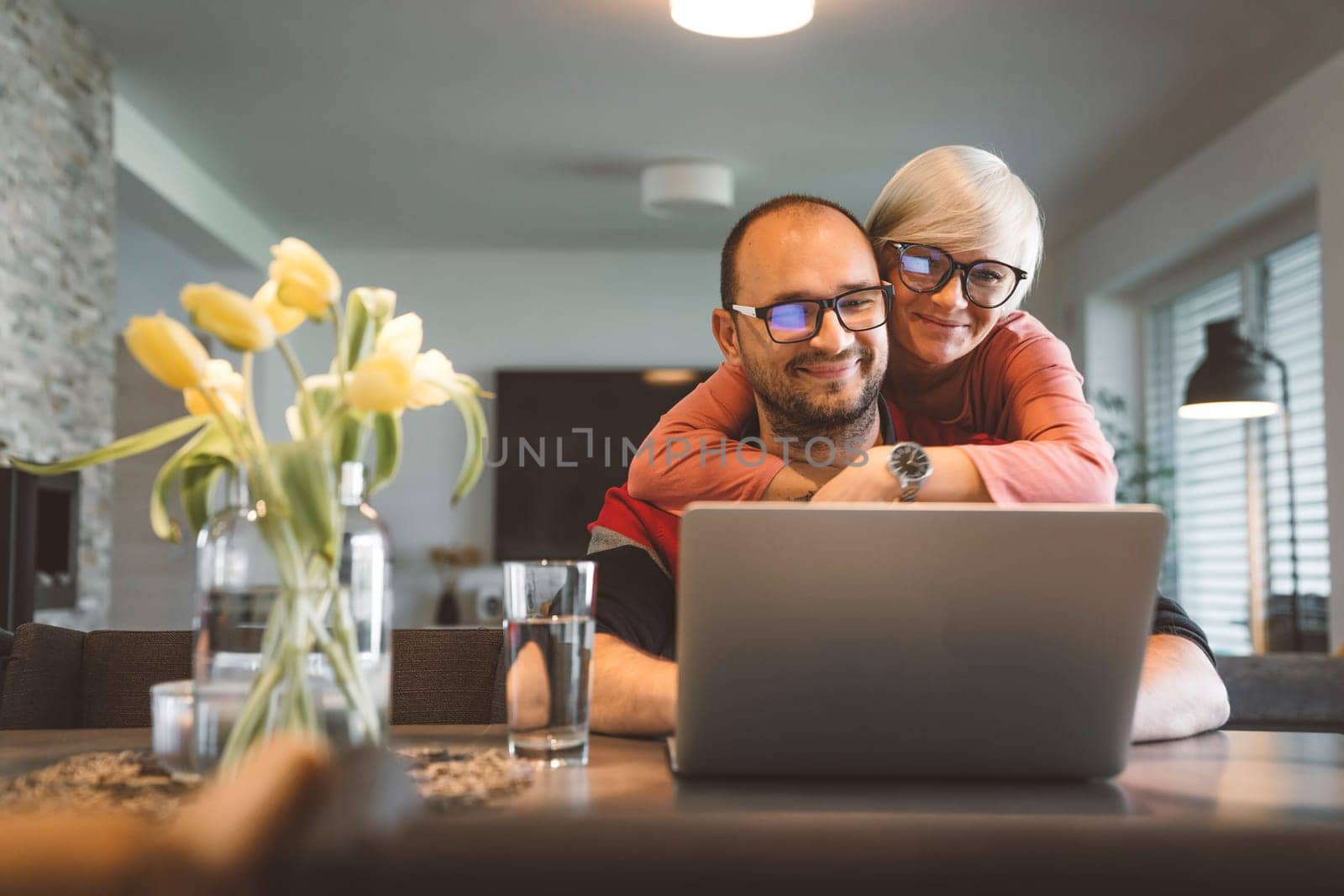 Wife hugging his husband while he works from home on the laptop by VisualProductions