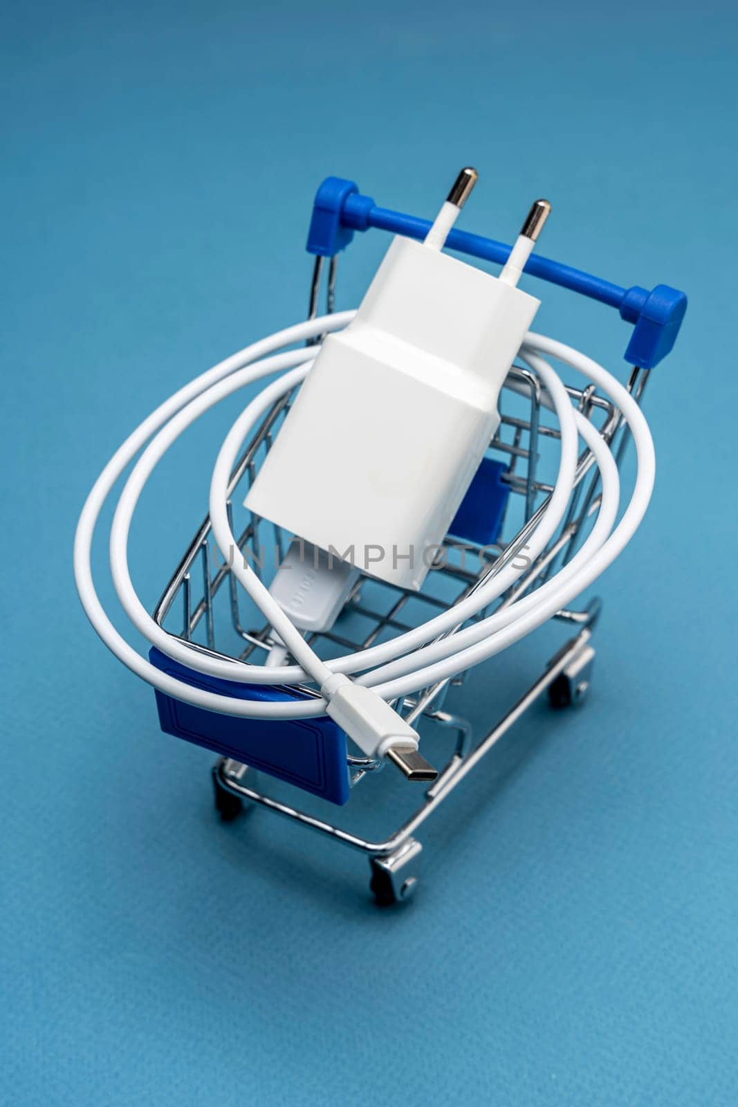 A phone charger in a supermarket cart on a blue background. Mini Shopping cart with Charger and Connecting Cable. vertical photo