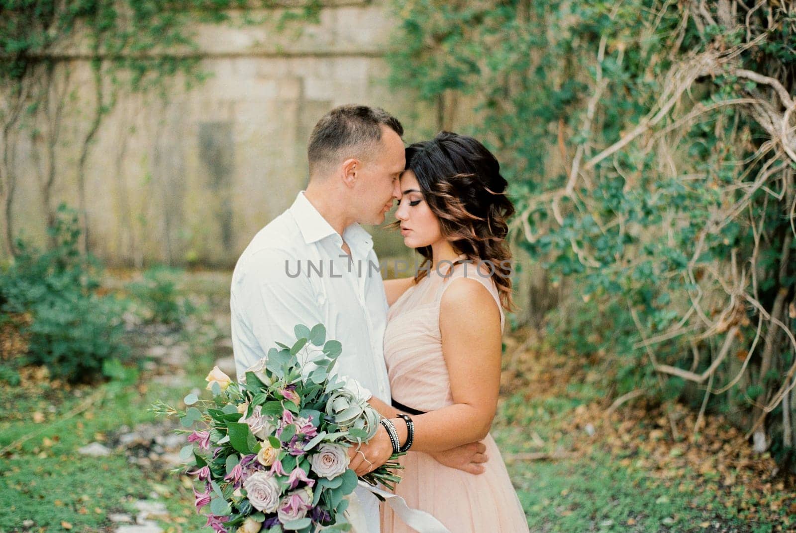 Groom hugs bride touching her forehead with his forehead near the ancient stone wall. High quality photo