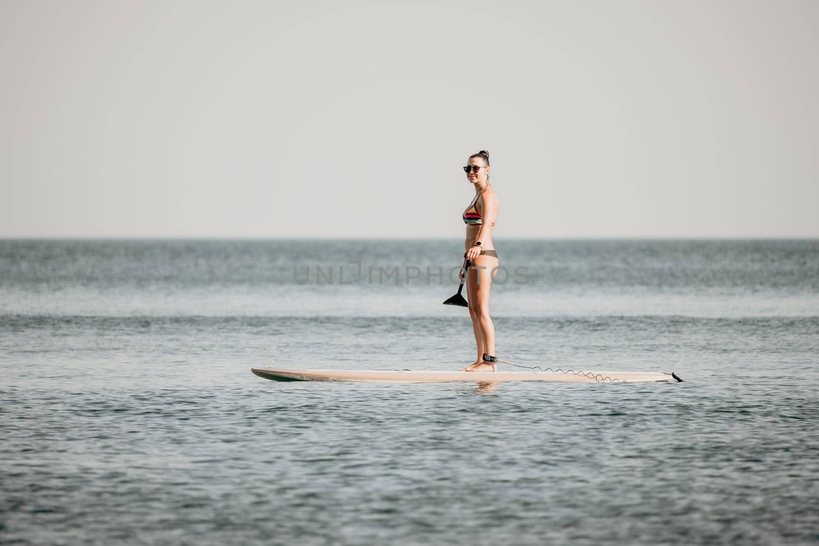 Sea woman sup. Silhouette of happy middle aged woman in rainbow bikini, surfing on SUP board, confident paddling through water surface. Idyllic sunset. Active lifestyle at sea or river