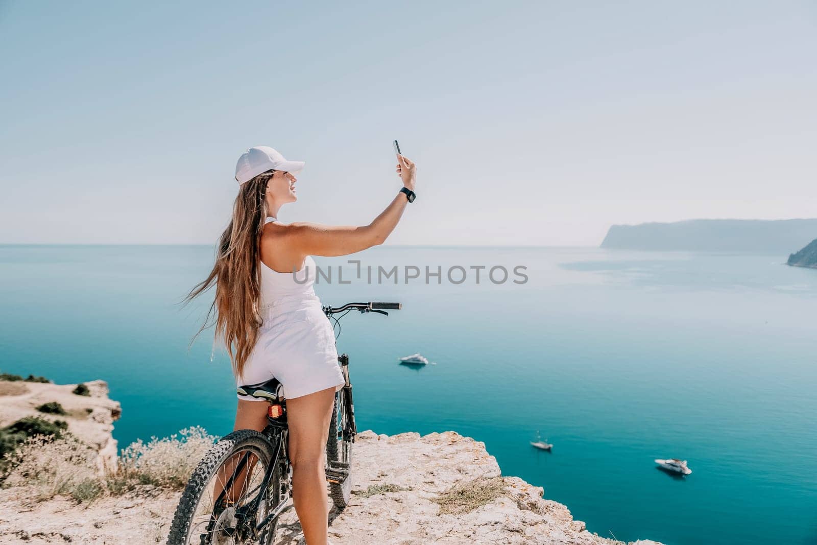 A woman cyclist on a mountain bike looking at the landscape of mountains and sea. Rear view of cyclist woman standing in front to the sea with outstretched arms. Freedom and healthy lifestyle concept