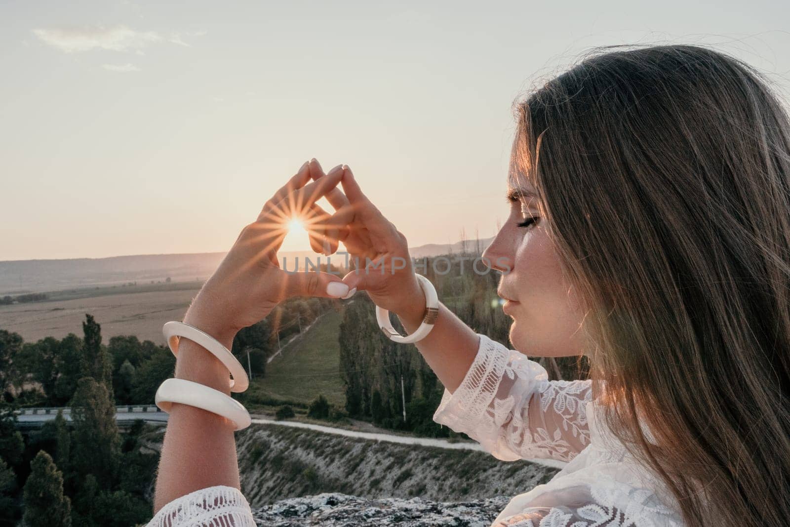 Happy woman in white boho dress making heart sign with hands on sunset in mountains. Romantic woman with long hair standing with her back on the sunset in nature in summer with open hands. by panophotograph