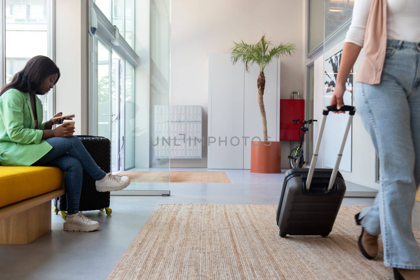 Hotel reception lobby. Female sitting on bench using mobile phone, woman walking by with trolley suitcase. Copy space. Lifestyle concept.