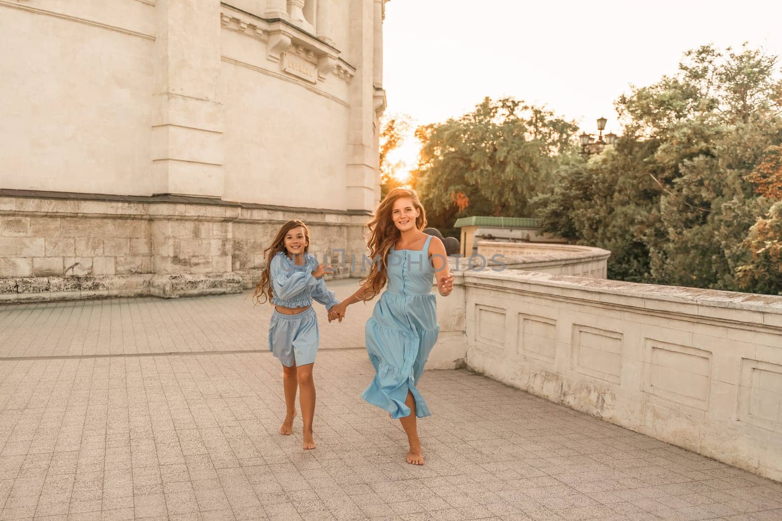 Daughter mother run holding hands. In blue dresses with flowing long hair against the backdrop of a sunset and a white building