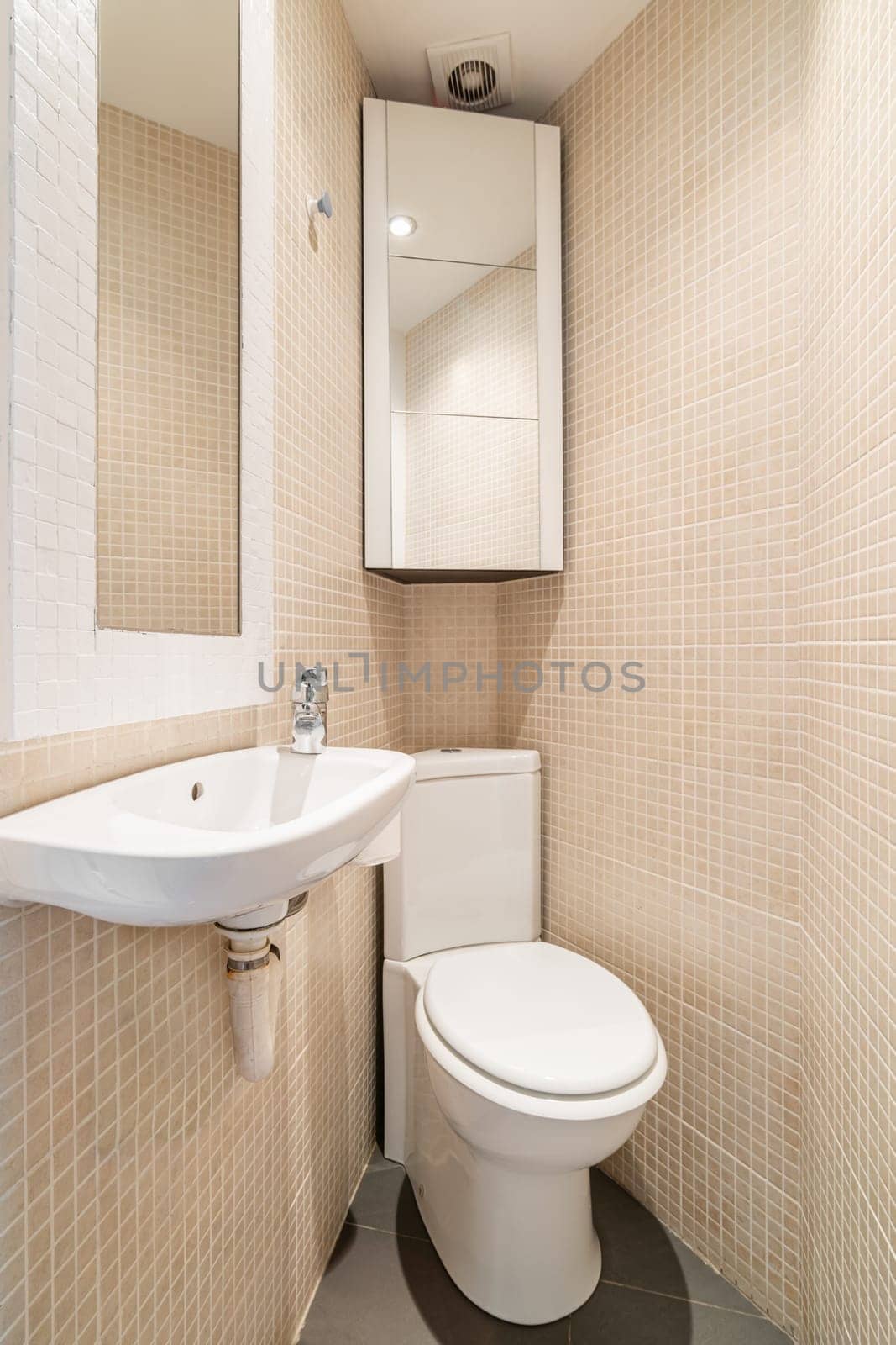 View of the toilet in the corner of compact tapered bathroom with beige mosaic tile, mirror, toilet and sink and wall cabinet. Concept of design solutions in a closed space.