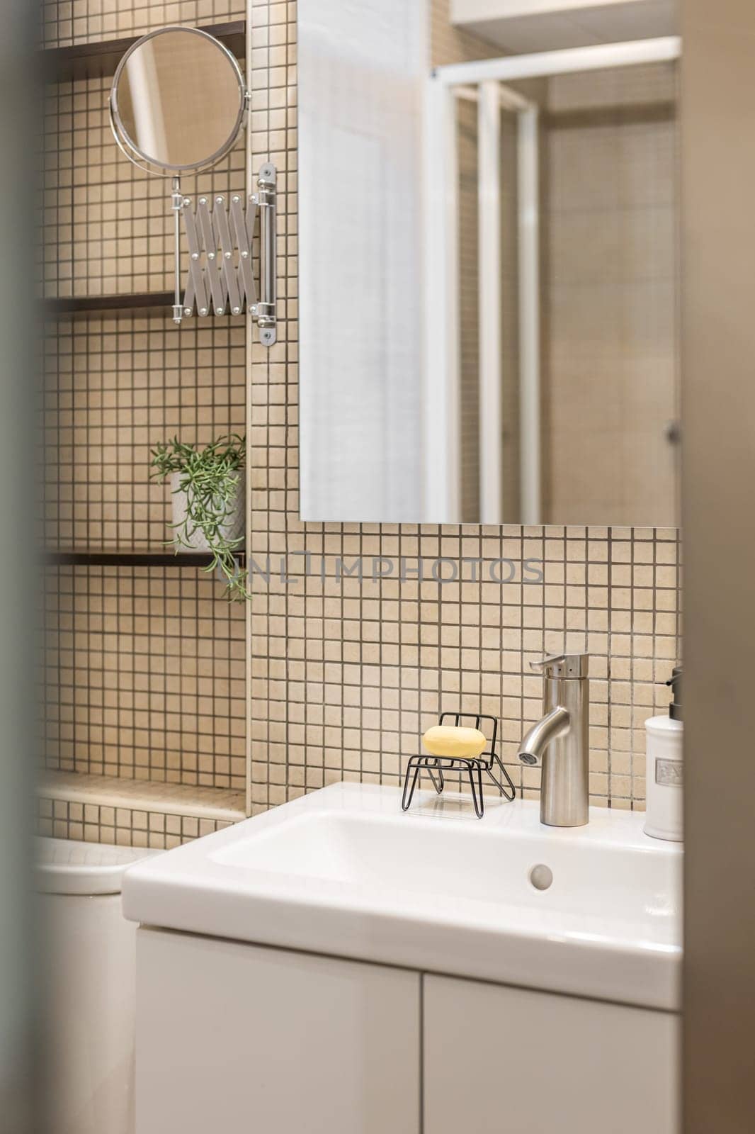 Close-up fragment of stylish bathroom with beige mosaic tiles, mirror, sink and shelves for things. Concept of a comfortable and usable bathroom in a small apartment.
