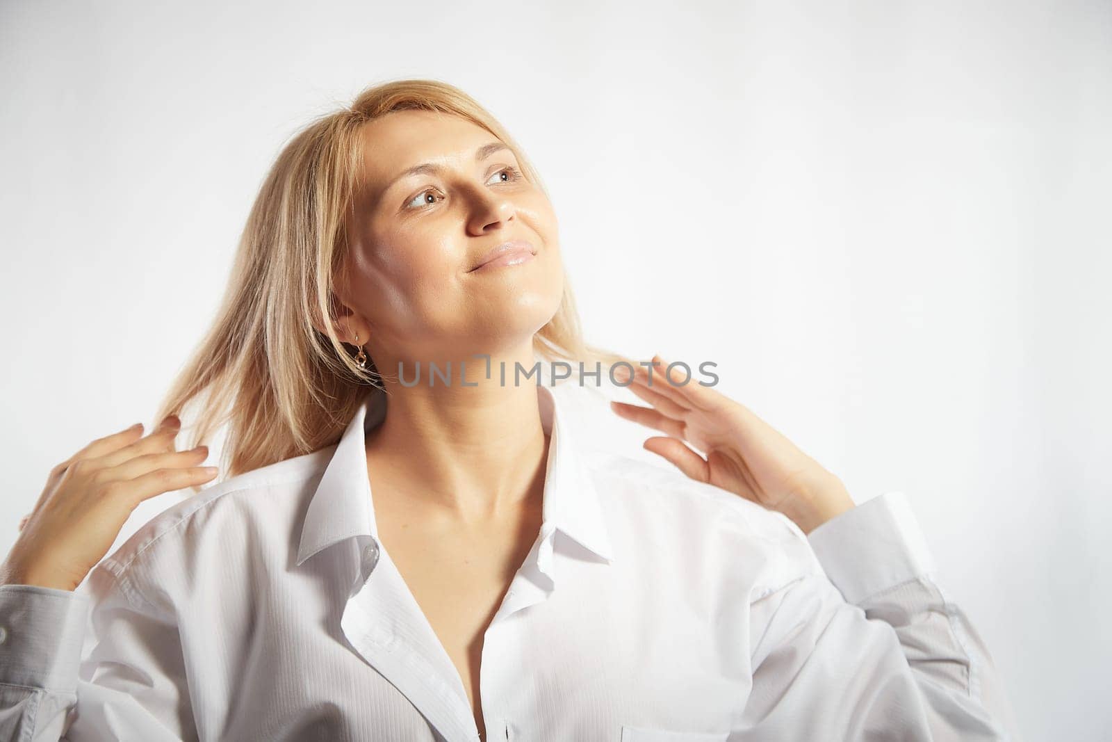 Portrait of a pretty blonde smiling woman posing on a white background and correcting her hair and hairstyle. Happy girl model in white shirt in studio by keleny