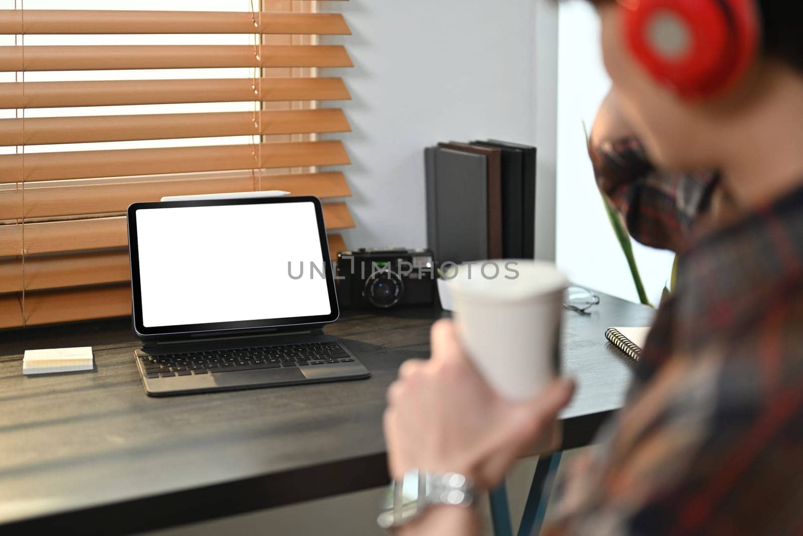 Cropped image of hipster man drinking coffee, sitting in front of digital tablet with blank screen on black wooden desk by prathanchorruangsak